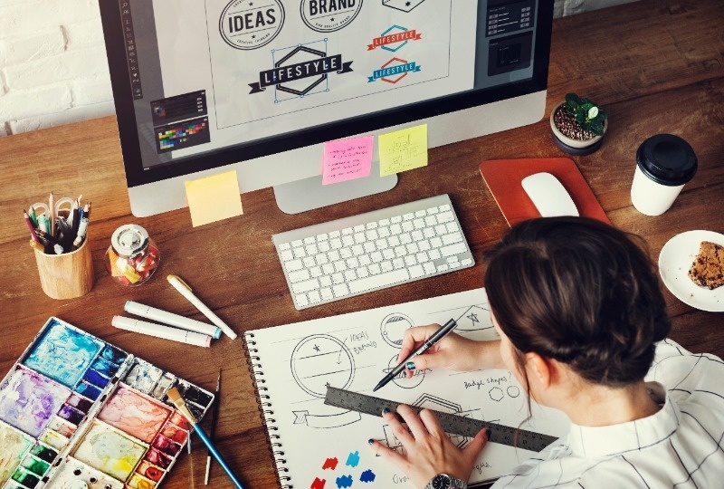 Graphic designer sitting in front of a laptop and sketching logos in a notepad